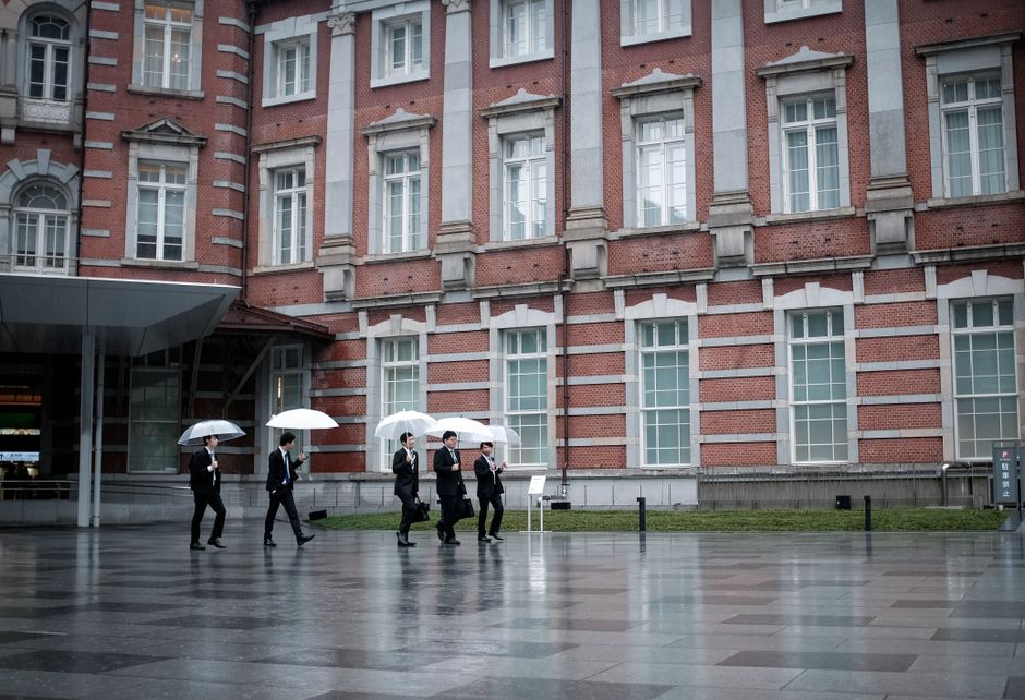 Businessmen in suits walking with umbrellas outside a historic building in Tokyo during a rainy day.